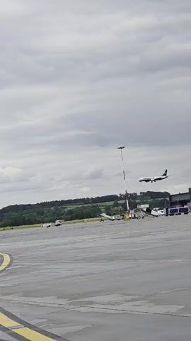 The video opens with a wide shot of an airport tarmac under a cloudy sky. In the foreground, a portion of the tarmac is visible with yellow and white markings. In the mid-ground, a white airplane with dark accents on its tail is in the process of landing or taking off. The plane is angled upwards, suggesting it is in the air. Further in the background, a line of trees forms a natural backdrop against the sky, with some buildings and a tall light pole also present. The tarmac appears wet, indicating recent rain. The overall atmosphere is overcast and muted.