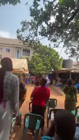 The video shows a group of people, mostly women, at what appears to be an outdoor event or gathering. The setting is a dirt yard with trees providing some shade. Several people are wearing colorful clothing, including dresses and head coverings. Some women are wearing red sashes. The women are gathered, talking, and some are dancing. There are chairs in the foreground, some stacked and others occupied.  A white tent structure is visible in the background, along with a building. The video is shot from a slightly low angle.