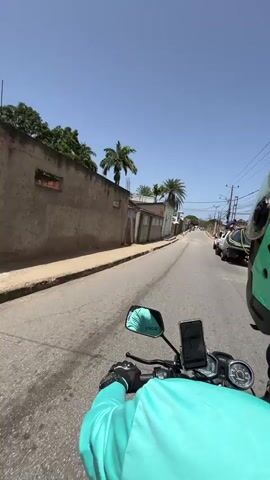 A first-person perspective video shows a person riding a motorcycle down a street in Los Cerritos, Venezuela, on a clear, sunny day. The rider, wearing a bright teal-green jacket and black gloves, has a phone mounted on the handlebars displaying a map. The motorcycle's mirror shows "Vickoo" text. On the right, a green helmet with "Rider" partially visible can be seen. The road appears paved but has some wear.

On both sides of the street, there are buildings with various architectural styles, including some with stone-faced walls and white facades. Numerous tall palm trees are scattered along the street. Utility poles with overhead wires line the right side of the road. In the distance, other vehicles are visible, suggesting a populated area. The sky is bright blue with no clouds.