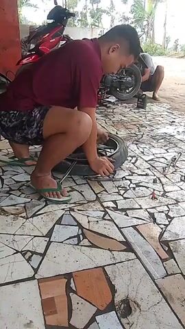 A person in a maroon shirt and patterned shorts crouches over a motorcycle tire on a floor tiled with irregular pieces of various colors. The person is using a tool, possibly a tire lever, to work on the tire. Another person is kneeling in the background, near a red motorcycle, and appears to be working on it as well. The environment looks like an outdoor repair area, with a dirt or gravel surface visible beyond the tiled area and some vegetation in the background. Tools and other objects are scattered around the workspace.
