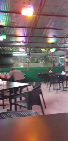 The video shows an outdoor dining area of a restaurant. There are multiple tables and chairs, with a corrugated metal roof overhead. Colorful lights are hung from the ceiling, casting a vibrant glow. Several people are visible, some seated at tables, and one woman is walking across the scene. The atmosphere appears casual and relaxed, possibly during the evening hours given the lighting.