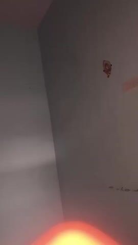 The video shows a close-up of a wall with some stains and a sticker. The wall is a light gray color and appears to be textured. There are several dark spots on the wall, which may be stains or dirt. In the upper right corner of the frame, there is a small, colorful sticker with what appears to be a cartoon character. A bright light source illuminates the left side of the wall, creating a strong contrast between the lit and unlit areas. The lighting is dim overall, and the focus is not sharp.