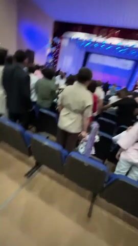 The video captures a group of people gathered in an indoor venue, likely an auditorium or conference hall. The initial seconds show a blurry, long-shot view of attendees seated in rows, with some standing. The lighting appears to be dim, with spotlights illuminating the stage area, which is out of focus. As the video progresses, the perspective shifts to a medium shot of a young woman with reddish hair in the foreground, taking a selfie or recording herself. Behind her, other attendees are visible, some standing and some seated. The overall atmosphere seems to be one of a formal event, possibly a ceremony, graduation, or a presentation, indicated by the attire of some individuals and the arrangement of the seating. The background reveals acoustic paneling on the walls and ceiling, typical of such venues.