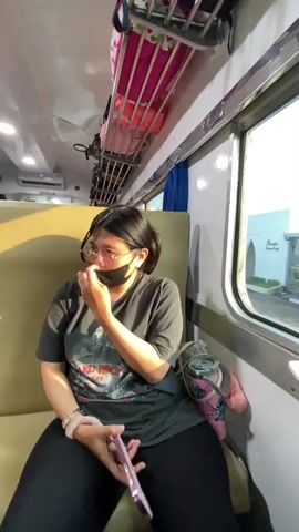 A woman wearing a black T-shirt, black leggings, glasses, and a black face mask is sitting on a light yellow train seat. She has her hair pulled back in a ponytail and is holding a pink cell phone in her left hand. The train car has a luggage rack above the seats and a window to the woman’s right. The window shows a building exterior with greenery. The woman turns to look at a small child who approaches her. The child, wearing a white T-shirt and dark pants, reaches out to touch the woman’s face.