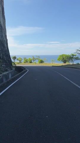 The video shows a black asphalt road curving along a cliffside. On one side of the road is a tall, rough-textured gray cliff face with some concrete retaining structures. The other side of the road features lush green trees and shrubs lining the edge of the cliff, overlooking a calm blue ocean. The sky above is bright blue with a few wispy white clouds. A white line marks the edge of the road. There are no people or vehicles visible in the frame.