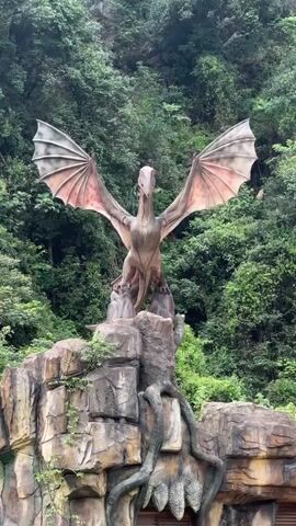 A statue of a dragon with outstretched wings stands on a rocky outcrop. The dragon is light brown and has large, leathery wings with pink undersides. It is positioned in front of a dense forest of green trees, with a rocky cliff face visible behind the trees. The statue appears to be made of stone or a similar material, and it is weathered and aged, with moss and vines growing on the rocks. The scene is well-lit, suggesting it is daytime, and the overall impression is one of ancient grandeur.