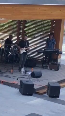 A band is performing on a raised wooden stage under a wooden overhang. Three musicians are visible. On the left, a person is playing drums. In the center, a man is playing a white electric guitar and singing. On the right, a person wearing a hat is playing a keyboard. Several speakers, microphones, cables, and amplifiers are on the stage. The background consists of a slatted wooden wall.