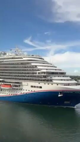 The video shows a large cruise ship, identified by the lettering as "Carnival Horizon," sailing on calm water under a bright, blue sky with scattered white clouds. The camera zooms in to focus on the ship's windows and balconies, where people can be seen inside or on the balconies, presumably passengers. The overall view is from the side, capturing the multi-deck structure of the ship with its white upper decks and a blue and red stripe along the lower part of the hull.