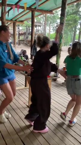 A group of people are dancing in a circle on a wooden platform under a covered structure. The structure is supported by wooden posts and has a green tarp overhead with colorful triangular flags strung across it. In the foreground, a woman in a turquoise hoodie and denim shorts is dancing. To her right, a person in a brown garment is also dancing. Further back, a woman in a patterned dress is leading a line of people in a dance. Behind her, a girl in a striped shirt and leggings is dancing. Other people are visible in the background, some sitting and some standing. The environment appears to be outdoors, with trees and some structures visible behind the covered area. There are tables with drinks and other items on them. The overall atmosphere seems festive and casual.