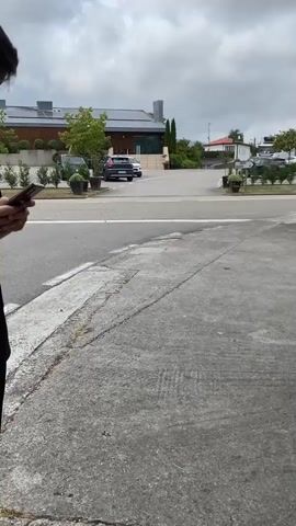 A person holding a phone films another person walking away from the camera. The person being filmed is wearing a black dress and appears to be pregnant. The person filming is wearing a white shirt and suspenders and is also holding a phone. They are walking on a paved surface with cars parked in the background. There are trees and buildings visible in the distance. The sky is cloudy.