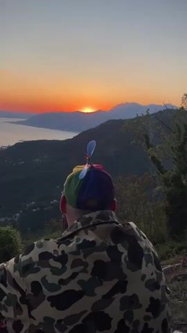 From behind, a man is looking out at a sunset view with distant mountains, a body of water, and forested hills. He wears a camouflage jacket and a colorful baseball cap with a spinning propeller. The sun is on the horizon. The propeller spins, stops, and spins again.