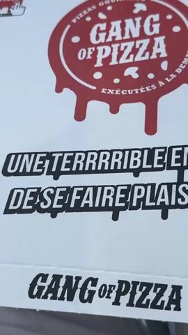 The video shows a close-up of a pizza box. The box is white and features a red logo with the words "GANG OF PIZZA" in a dripping font. Below the logo, text reads "UNE TERRRRRIBLE ENVIE DE SE FAIRE PLAAAISR?". There are also cartoonish bullet hole graphics on the box. On the side of the box, the word "HO" is handwritten in black marker. The pizza box is resting on a white surface, possibly fabric, and a person's hand with a ring is visible in the lower portion of the frame. In the background, a red object and some wires are visible, suggesting the box is in a car or similar environment. The video also shows a bottle with a blue cap peeking out from under the box.