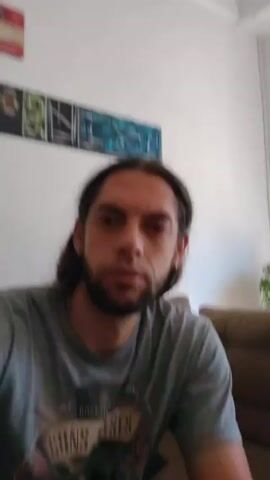 A man with long, dark hair, a beard, and a grey Guns N' Roses t-shirt is sitting on a brown couch indoors. He takes a bite of something in his hand, chews, and swallows. Behind him is a white wall with a multi-panel abstract painting and a houseplant. The man is facing the camera.