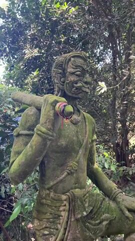 The video shows a weathered stone statue of a grotesque figure, covered in moss. The statue is in a lush, green environment, likely a jungle or a garden, with trees and foliage visible in the background. The statue is holding a cylindrical object over its shoulder. At the end of the video, a small lizard is perched on the head of the statue. The lighting suggests it is daytime.