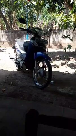 The video shows a blue motorcycle parked outdoors. The motorcycle is positioned on a dirt ground, and it is partially shaded by trees and leaves overhead. Behind the motorcycle, there is a wall and some sparse vegetation. The lighting suggests it is daytime. The camera appears to be handheld and is moving slightly.