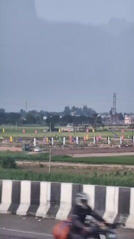 The video shows an outdoor scene, likely in a rural or semi-urban area. In the foreground, a concrete barrier with black and white stripes is visible, suggesting the perspective is from a road or highway. Beyond the barrier, a field is seen with rows of flags or banners displayed on poles. These flags are multicolored and appear to be part of some sort of setup or event. In the background, there are green fields, some buildings, trees, and a tall industrial tower or antenna. The sky is overcast with a hazy or foggy appearance, obscuring the distant landscape. A white car is parked in the field near the flags. The overall atmosphere is muted and somewhat gloomy due to the weather conditions.