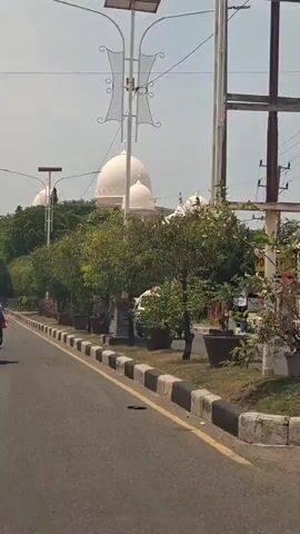 The video shows a road with a sidewalk lined with potted plants and trees. A large white domed building, possibly a mosque, is visible in the background. Several motorcycles are seen on the road, with one rider prominently featured at the beginning of the clip. Overhead power lines and decorative streetlights are also present. The sky is clear and the sun appears to be shining. The overall scene suggests a daytime view of a moderately busy urban or suburban environment.