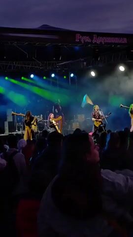 A live band performs on a dimly lit outdoor stage under a twilight sky. The band members, dressed in what appear to be medieval-inspired or fantasy costumes, play various instruments including guitars, a harp, and possibly a flute. One member is singing into a microphone. Green and blue stage lights illuminate the performers and create beams of light that cut through the smoky atmosphere. In the background, the words "Русь Дружинная" are visible on a banner above the stage. A large crowd of spectators is gathered in front of the stage, their silhouettes visible against the lights. The overall scene suggests an outdoor music festival or concert.