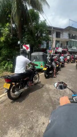 The scene is shot from the first-person perspective of someone sitting on a motor scooter in traffic on a paved road in a tropical environment. Numerous other motorcycles and a three-wheeled vehicle (auto rickshaw) are also stopped in the traffic. The sky is partly cloudy. The other vehicles are in front of the camera. There are two- and three-story buildings visible, and several people walking around on the sidewalks. The traffic is heavy. A sign showing a right turn is visible at the edge of the road. The first person appears to be getting ready to move forward when the traffic clears.