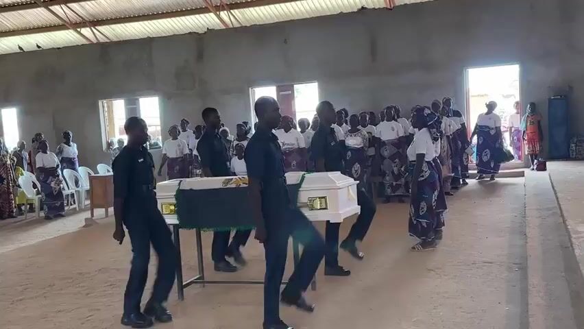 This video captures a solemn funeral service. In the foreground, several men in dark uniforms are marching in formation, stepping in unison, with their left legs raised high. They are escorting a white casket, draped with a green cloth and accented with yellow trim, which rests on a table. Behind the marching pallbearers, a gathering of people, many wearing white shirts and patterned skirts, are observing the proceedings. The environment appears to be a simple hall with concrete walls, a corrugated metal roof, and natural light filtering through windows. Further in the background, other individuals are visible, some seated and others standing. The scene conveys a sense of respect and ceremony for the deceased.