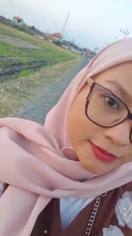 A selfie video is taken outdoors in daylight. The camera is focused on the face of a young woman wearing a light pink hijab and glasses. She is looking at the camera and smiling. In the background, there is a road and some fields. The video is short and the woman's face is the main focus.