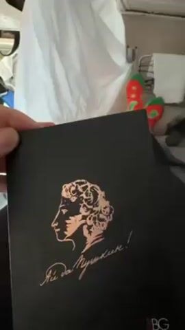 Here is a description of the video:

The video shows a hand holding up a black box with a bronze bust of Alexander Pushkin on the front. The text below the image is in a stylized font, likely in Russian, and ends with an exclamation point. The lower right corner of the box features the initials "RBG" in red. The box is then opened, revealing a makeup palette with four shades: a brown contour, a pink blush, and two highlighting shades. In the background, there is a white sheet or curtain, along with a pair of novelty socks with green stems and red fruits on them. The socks are worn by someone in the background, as only their legs and feet are visible. The environment appears to be a cluttered space, possibly a bus or vehicle interior.