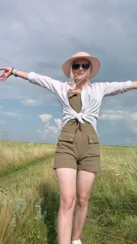 A young woman with blonde hair and sunglasses stands in a grassy field wearing a white shirt, green overalls, and a straw hat. She is barefoot and raises her arms above her head. The sky is cloudy and blue. She turns her back to the camera and then faces it again.