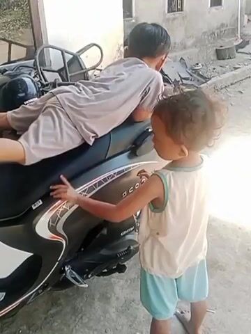 Two young children are interacting with a parked scooter outside a building. One child, wearing a light-colored t-shirt and light blue shorts, stands to the right of the scooter, facing it and the other child. They have dark, curly hair and are looking towards the other child. Their right hand is held up, with the index and middle fingers extended, forming a peace sign. The other child, wearing a beige t-shirt and beige shorts, is seated on the scooter's seat, facing away from the camera and towards the building. They are leaning forward, with their arms resting on the scooter's seat. Their legs are visible, extending over the side of the scooter. The scooter is black with white and red accents. The environment appears to be an outdoor, possibly urban or semi-rural setting, with a concrete ground and a building visible in the background. The lighting suggests it is daytime.