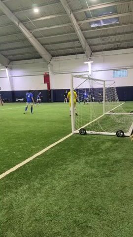 A group of people are playing soccer indoors on an artificial turf field. The field is green with white lines. There is a soccer goal with a net and wheels on the right side of the frame. A person in a yellow goalkeeper jersey with the number 29 on the back is standing in front of the goal. Several other people in blue jerseys are on the field, some of whom appear to be playing. The ceiling of the building is visible, with exposed beams and lighting fixtures. The walls of the building are light-colored. The overall environment suggests a sports facility.