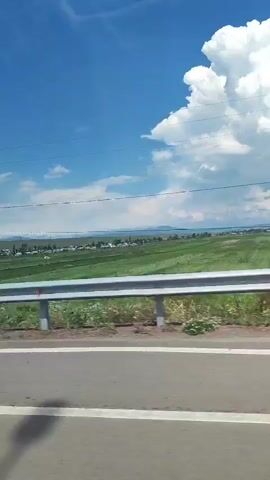 This video captures a scenic view from a moving vehicle, likely a car, on a bright, sunny day. The sky is a vibrant blue with fluffy white clouds scattered across it, suggesting pleasant weather. The scene depicts a rural or semi-rural landscape. In the foreground, a grey asphalt road is visible with a white line marking its edge. A silver metal guardrail runs along the side of the road, separating it from a grassy verge. Beyond the guardrail, a vast expanse of green fields stretches into the distance, hinting at agricultural activity.

In the middle ground, a small town or village is nestled amongst the fields, characterized by scattered buildings and trees. A prominent feature in the landscape is a tall, grey utility pole with wires stretching across the frame, indicating the presence of electricity infrastructure. Further into the distance, the terrain appears to rise into rolling hills or mountains.

The video seems to be shot from a side window of the vehicle, offering a panoramic view of the surroundings. There are no people clearly visible in the frame, and the activity taking place is simply the motion of the vehicle through the landscape. The time of day appears to be daytime, possibly late morning or early afternoon, given the bright sunlight. The overall impression is one of a peaceful journey through a picturesque countryside. No discernible text is visible in the video.