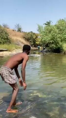 A young, lean, shirtless Black man stands in a shallow, clear river on a sunny day. He wears checkered boxer shorts. He looks around, appearing to admire the scenery. He touches the back of his head with one hand and then looks down at the water. The environment includes low trees, bushes, and some rocks on the bank. Another individual is faintly visible further along the river. The sky is clear and blue.