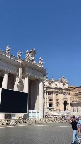The video shows a sunny, daytime view of St. Peter's Square in Vatican City. The sky is clear blue. In the foreground, people are walking across the square, including one man wearing a straw hat and another with a backpack in the style of the British flag. To the left, there's a large video screen and a low, wooden fence. The dominant features are the architecture of the Vatican buildings, with detailed stonework, statues atop the structures, and rows of columns. The video pans slightly to the right, showing more of the architecture and the crowd of people in the square.