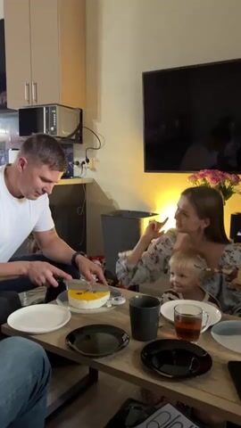 A man in a white t-shirt is cutting a cake on a wooden table. He is on the left side of the frame, and the cake is in front of him. The cake is round, with a yellow topping and some decorations on top. The man is holding a knife and is in the process of slicing the cake.

To the right of the man, a woman in a floral dress is holding a young child on her lap. The woman is smiling and looking at the man, and she seems to be interacting with the child. The child is also looking towards the cake.

On the table, there are several plates, cups, and a tea set. The plates are white and black, and the cups are dark gray. There is also a glass of tea. In the background, there is a television screen, a cabinet, and a window. The scene appears to be indoors, in a home setting. The lighting suggests it could be evening or a dimly lit room.

The overall mood of the scene is warm and celebratory, suggesting a family gathering or a birthday party. The presence of the cake and the happy expressions of the people indicate a positive and joyful occasion. The man's focused action of cutting the cake adds to the sense of a special moment being shared.