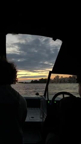 The video shows a view from inside a boat looking forward. Two silhouetted figures are visible, one in the foreground looking towards the left and another in the right side of the frame, appearing to be steering the boat. The boat is moving on a body of water, with ripples visible on the surface. In the distance, a shoreline with trees and lights is visible. The sky is cloudy with a strip of orange and yellow near the horizon, indicating either sunrise or sunset. The overall lighting is dim.