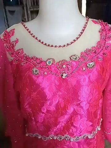 The video displays a close-up of a bright pink, lace dress with intricate beading and jewel embellishments around the neckline and waist. The dress is on a mannequin, and a hand briefly enters the frame to adjust it. The background appears to be an indoor setting, possibly a workshop or store, with blurred objects and lighting visible. The focus is entirely on the detailed craftsmanship of the garment.