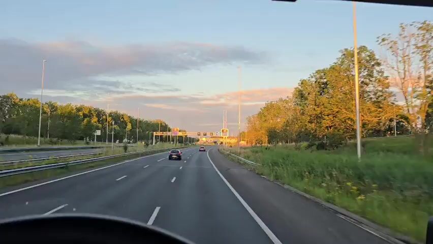 This video shows a first-person perspective from inside a vehicle driving on a highway at sunset. The road has multiple lanes and is bordered by trees and grassy areas. There are a few cars visible ahead on the highway, and overhead signs are present. The sky is clear with some clouds, illuminated by the setting sun, casting a warm glow on the trees. The steering wheel of the vehicle is partially visible in the foreground.