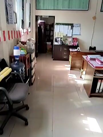 The video shows an indoor office space. The camera is panning slowly from left to right. On the left wall, there are windows covered by white curtains and red and white striped decorations. Below the windows, there is a black office chair, a multi-drawer cabinet, and a water cooler. Further to the right, there is a hallway leading to another room. On the right side of the hallway, there is a dark brown cabinet with drawers and shelves, with papers and other items on top. In front of this cabinet, there is a wooden desk with papers. To the right of this desk, there is another wooden desk with files and papers. The walls are painted a light color, and the floor is tiled. A large chart with a lot of text is mounted on the wall above the desks. Another smaller chart is mounted on the wall to the right. The lighting suggests it is daytime, with sunlight casting shadows on the floor. The space appears to be an office with multiple workstations and storage areas.