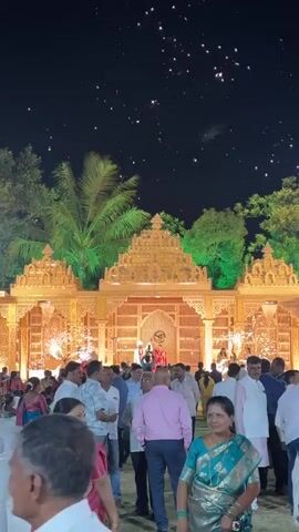 The video shows a large gathering of people at an outdoor wedding reception at night. They are standing in front of a large, ornately decorated stage-like structure lit with warm, golden light. The structure has a pyramid-like top and a wooden screen with geometric patterns. Behind the structure are palm trees and other trees lit with green lights. Two people, presumably the bride and groom, are standing on the structure, visible through the screen. They are waving to the crowd. Sparks are shooting up on either side of the structure. Most of the people in the crowd are facing the structure, looking at the bride and groom. They are wearing a variety of clothing, including traditional Indian clothing like sarees and western attire such as shirts and trousers. Some people are taking photos or videos with their phones. The night sky is visible, with a few small, indistinct lights scattered across it.
