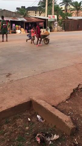The video shows a daytime scene in what appears to be a developing country. In the foreground, there is a concrete curb with litter scattered around it. Further away, three children are pushing a wheelbarrow with a large object inside. To the left, an adult female stands near a small shop. In the background, there are buildings and trees. A POS advertisement sign is visible. The overall impression is one of a busy but somewhat rundown street scene.