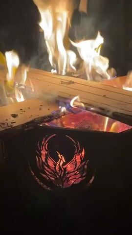 The video captures a close-up view of a metal fire pit at night, illuminating its surroundings with bright, dancing flames. Logs of wood are burning intensely within the pit, producing vibrant orange and yellow flames that reach upwards, accompanied by wisps of smoke. The fire pit itself is dark-colored and features a striking cutout design on its side: a phoenix with outstretched wings, glowing fiery red and orange from the embers and flames inside. The internal glow of the fire is prominently visible through the intricate details of the phoenix, creating a captivating visual effect. No people are visible in the frame, but the ambient sounds include human voices speaking in what appears to be Russian, along with the distinct crackling of the burning wood, suggesting a social gathering around the fire. The overall atmosphere conveyed is one of warmth and an outdoor evening setting.