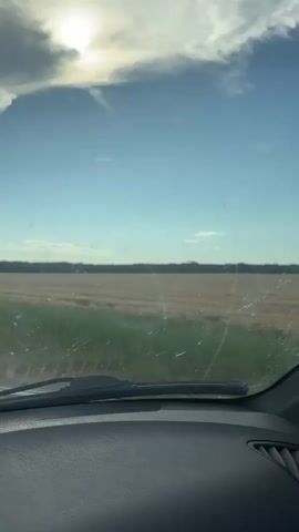 The video shows a view from inside a vehicle looking out through the windshield. The windshield has some streaks or dirt on it. Outside, a clear blue sky with the sun visible in the upper left corner is seen. Below the sky, a wide, flat field stretches to the horizon. The field appears to be a harvested crop, possibly wheat or hay, with a lighter brown color. A line of green grass or vegetation runs along the edge of the field closest to the vehicle. The interior of the vehicle is partially visible, including the dashboard. The overall impression is a view of a rural landscape during daylight.
