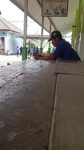 A person wearing a blue shirt and a black cap is leaning on a wooden table, looking at their phone. They are in an outdoor area with a covered walkway. In the background, several people in blue shirts, possibly students or staff, are walking. There is a building with a light-colored wall and a tiled roof on the left side of the frame. A poster is hanging on the wall of the covered walkway. The weather appears to be overcast or shaded.