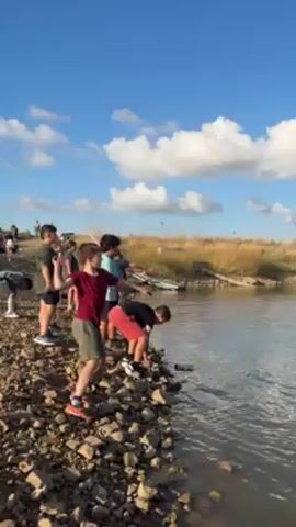 A group of approximately 15 people, mostly children and teenagers, are gathered at the edge of a body of water. They are on a rocky shore, picking up stones and throwing them into the water. Some are standing further back, observing. The sky is blue with scattered white clouds. The opposite bank is grassy and slopes upward. A ladder is lying horizontally on the grass. The atmosphere appears casual and playful.