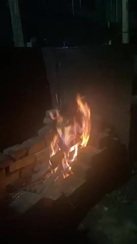 The video shows a close-up of a small bonfire at night. The fire is burning in a makeshift fire pit constructed from bricks. The flames are a mix of yellow, orange, and purple, indicating different temperatures. The surrounding area is dark, with only the light from the fire illuminating the scene. There are no people visible in the video. The fire appears to be made of wood.