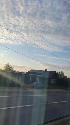 The video shows a rural landscape with a road in the foreground. A few houses and trees are visible in the background. The sky is blue with scattered white clouds. There is a billboard with an advertisement on it, and a lamppost. The sun is shining, casting long shadows. The video appears to be filmed from a moving vehicle, as evidenced by the blur of the passing scenery and the road markings.