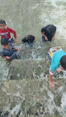 A group of five children are playing in water that appears to be flowing over concrete steps. One child in a red shirt is on the left side of the frame, partially submerged. Next to him, another child in a black shirt is also partially submerged, looking down. Further to the right, another child in a black shirt is kneeling in the water. In the center, a child in a grey shirt is sitting on a step, looking towards the camera and smiling. To the far right, a child wearing a colorful shirt is partially visible, with their legs in the water. The water is choppy and appears to be moving. The steps are made of concrete and are worn by the water.