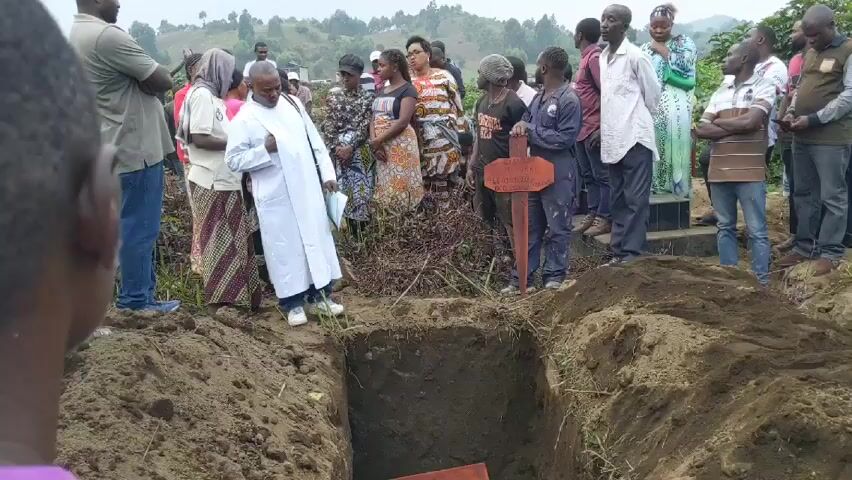 Here is a forensic description of the scene:

The video shows a funeral. A group of people, mostly appearing to be African, stand around a grave. A man in a white robe is present. There is a wooden cross with text on it near the grave. The landscape appears to be rural and hilly. The sky is overcast. The people are somber and respectfully standing. The video seems to be taken from the perspective of someone at the edge of the grave site.