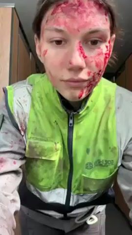 A woman in a gray and bright green work uniform stands in a narrow restroom with beige stall doors. She has what appears to be fake blood smeared on her face. She initially looks at the camera, then turns slightly to the side. Then, she puts her hands in her pockets and makes a peace sign with one hand. The uniform and the surrounding restroom also have blood-like stains on them.