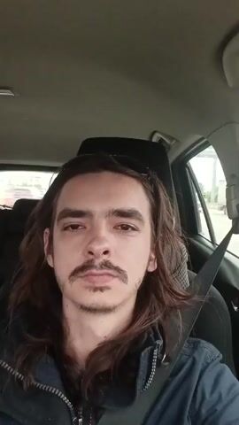A man with long brown hair and a mustache sits in the driver's seat of a car, looking directly at the camera. He is wearing a dark blue jacket with a zipper. A black seatbelt is visible across his chest. The interior of the car is visible, including the headrest and part of the car's roof. Outside the car window, other vehicles and buildings are visible. The video appears to be a selfie taken while driving.