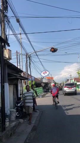 The video captures a street scene with a person riding a bicycle away from the camera. The cyclist is wearing a red and blue shirt and black shorts, with a red cap. The road is paved, and there are buildings and shops on the left side. Overhead, a complex network of electrical wires is visible against a clear blue sky. A traffic light with a yellow signal is hanging over the intersection. On the sidewalk to the left, a person wearing a striped shirt and shorts stands facing the camera. Another person in a high-visibility vest stands near the cyclist, holding a red and white sign. Cars and other vehicles are present on the road in the distance. A sign for "PLANET" is mounted on an oval-shaped structure above the shops.